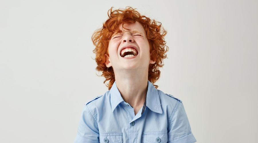 Close up of joyful little kid with ginger curly hair and freckles in blue shirt laughing hard watching comedy with parents in cinema